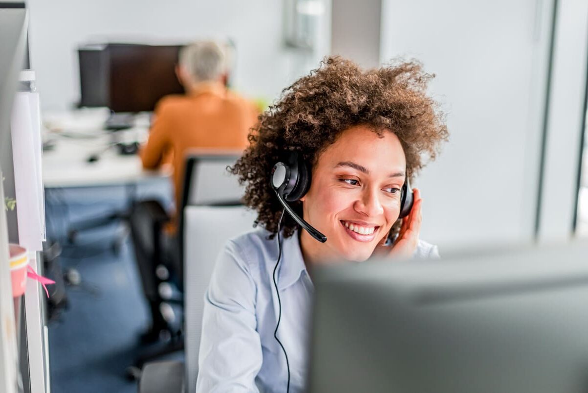 Photo of smiling, Cheerful mixed-race girl call working in center operator, wearing business attire. Young Customer service executive woman working at office. Female Customer service executive talking on headset in office. Young attractive friendly customer service answering customer questions on the telephone with toothy smile.