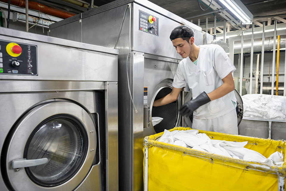 Focused man working at an industrial laundry service loading washing machine wearing protective gloves - Business industry concepts
